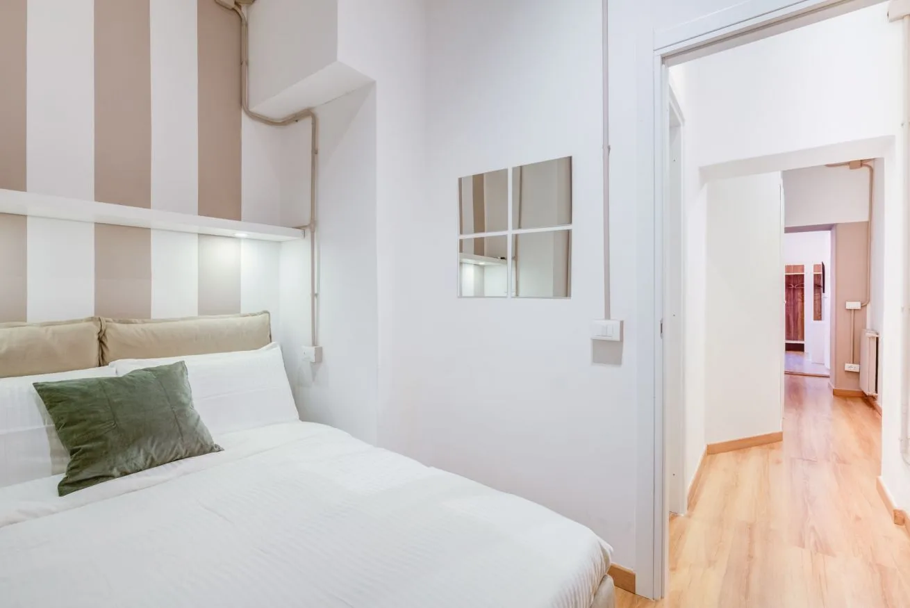 A bright bedroom with a neatly made bed and an open doorway to a hallway.