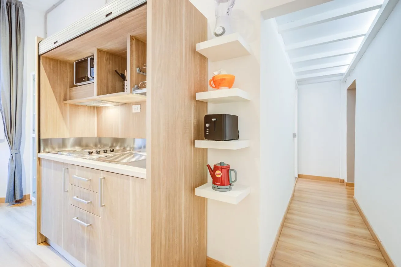 A compact kitchenette with wood cabinetry sits beside a bright hallway.