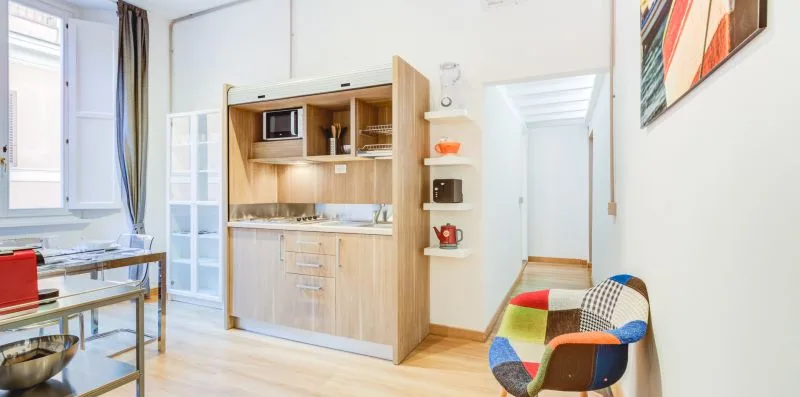 Una pequeña cocina luminosa con gabinetes de madera clara y una silla colorida.