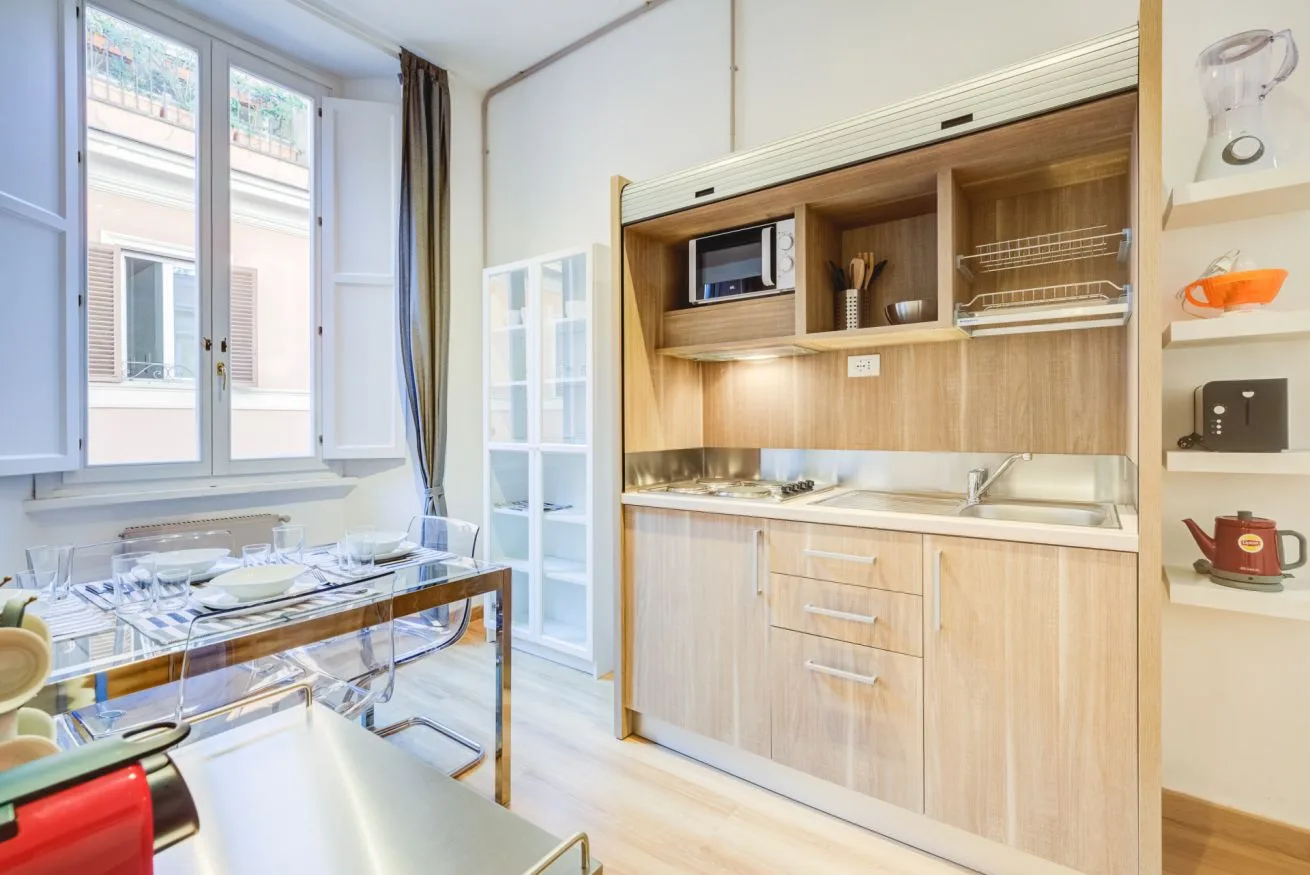A bright dining area with a glass table beside a wood-front kitchenette.