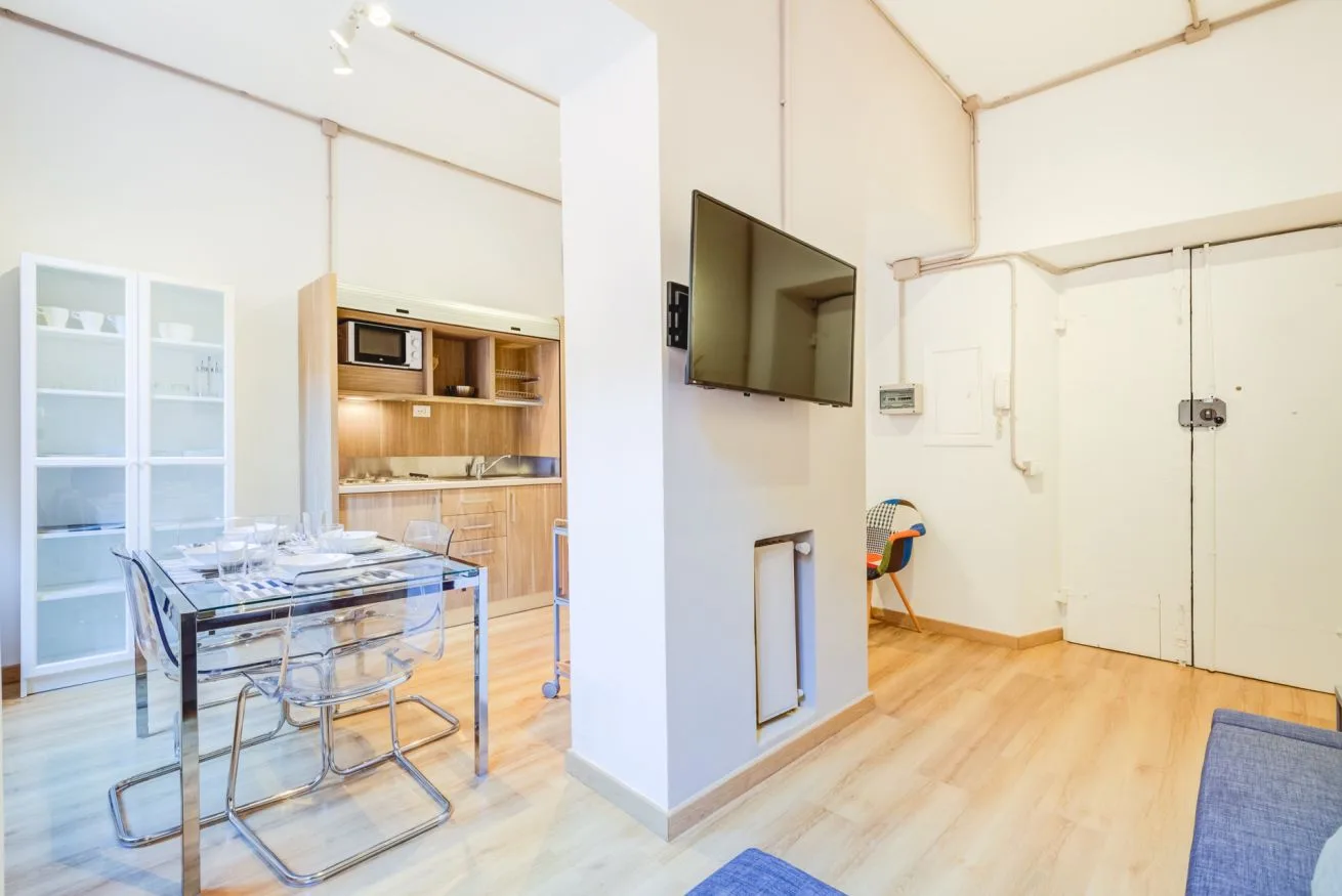 A bright open-plan room with a dining table, kitchenette, and wall-mounted television.