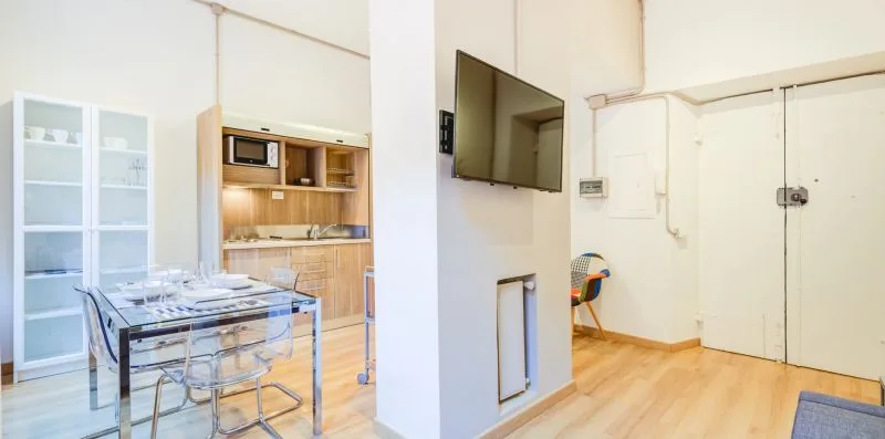 Una estancia luminosa con mesa de comedor, cocina pequeña y televisor en la pared.