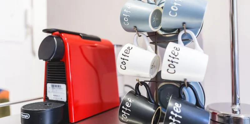 A red coffee machine sits beside a rack of hanging mugs.