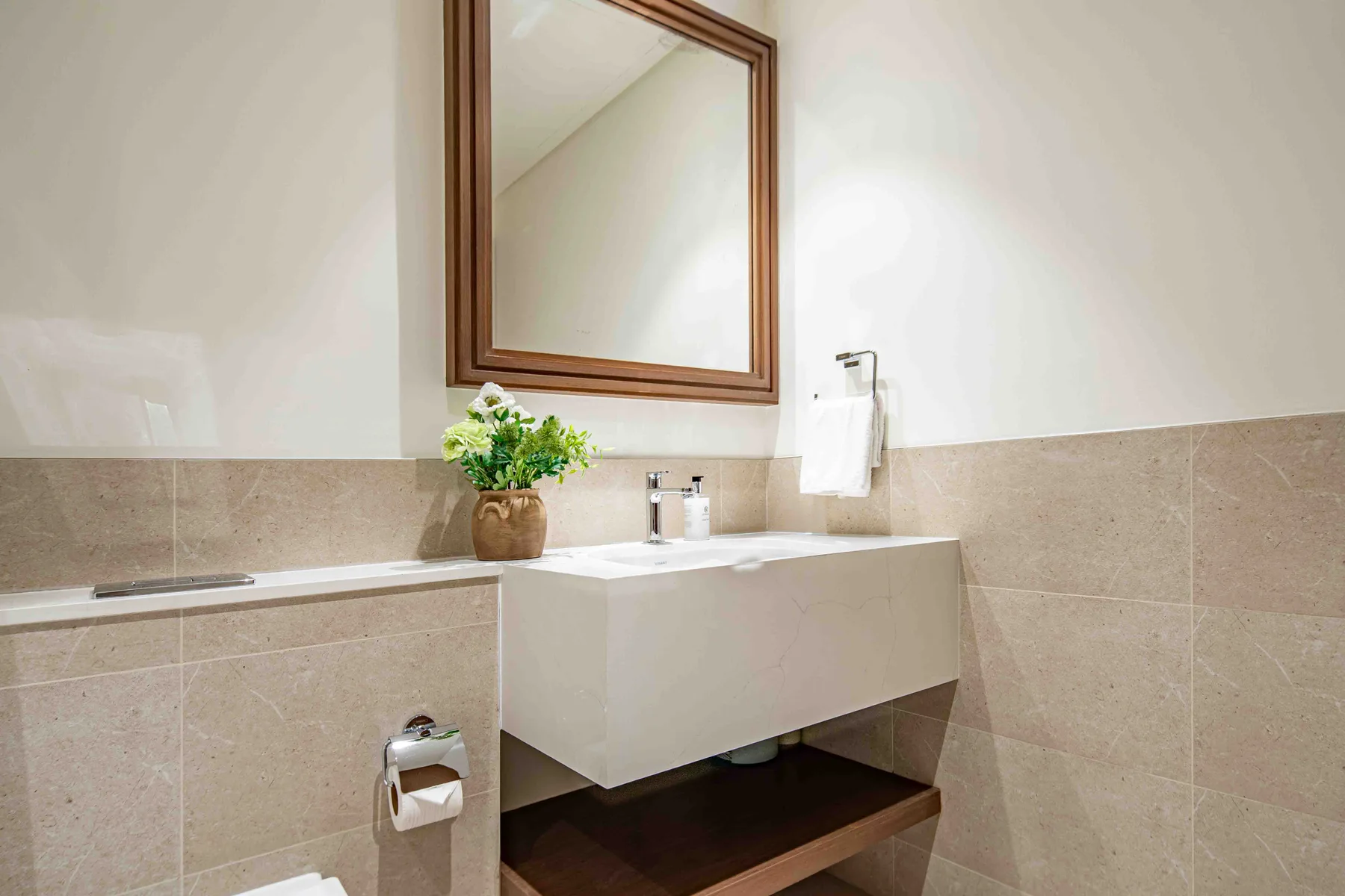 Un baño con lavabo suspendido, espejo y un pequeño arreglo floral.