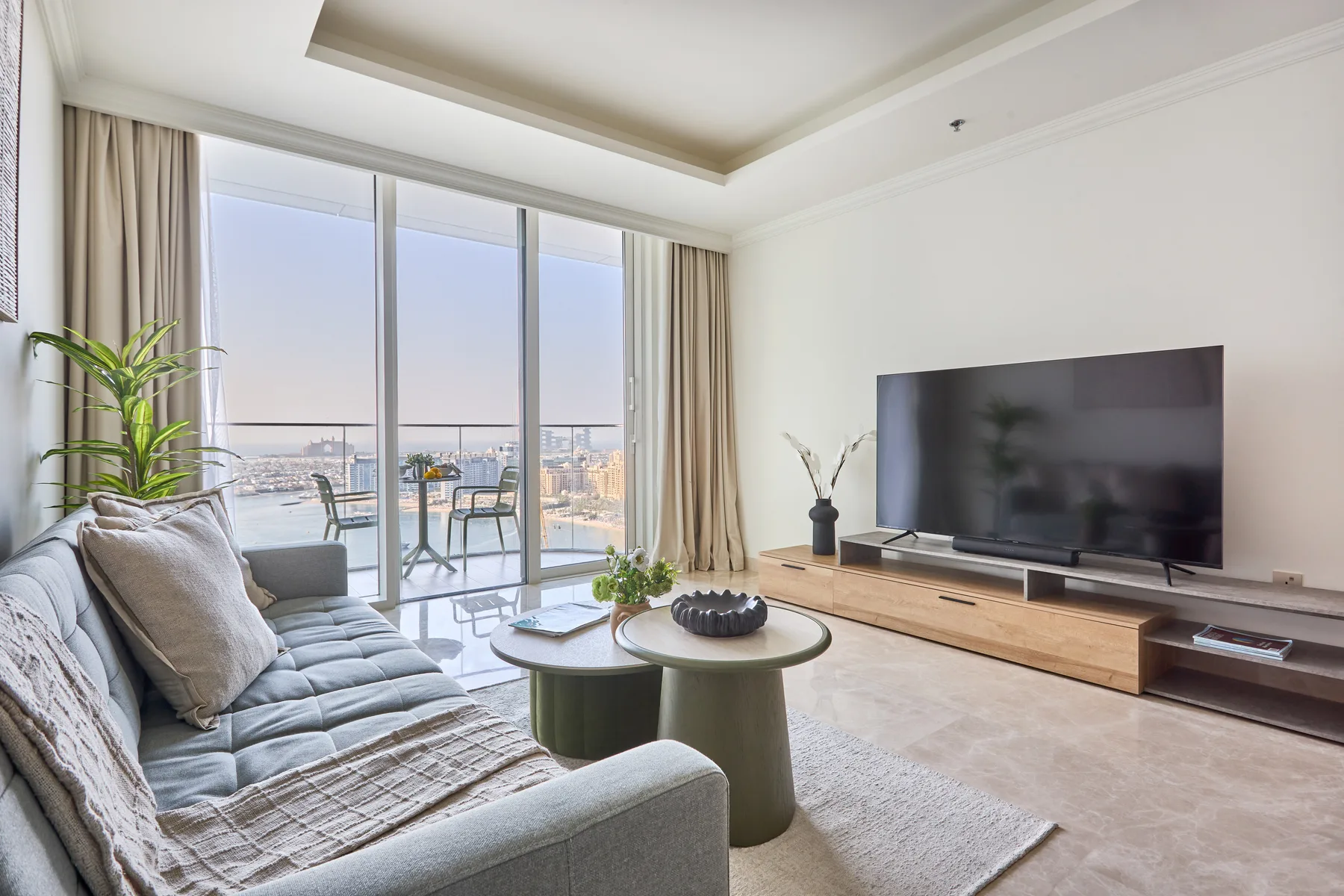 Sala de estar con sofá, mesa de centro y televisor frente a grandes ventanales.