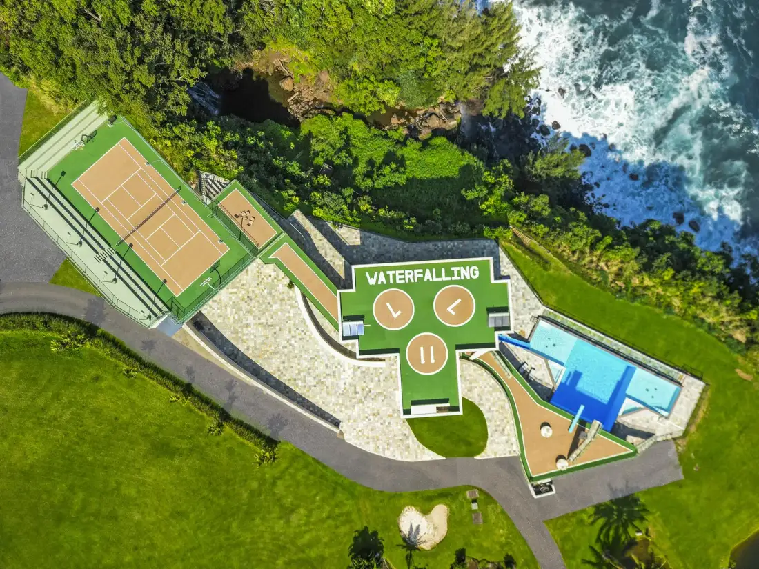 An aerial view shows outdoor tennis courts, a waterfalling sign, and a pool beside the coast.