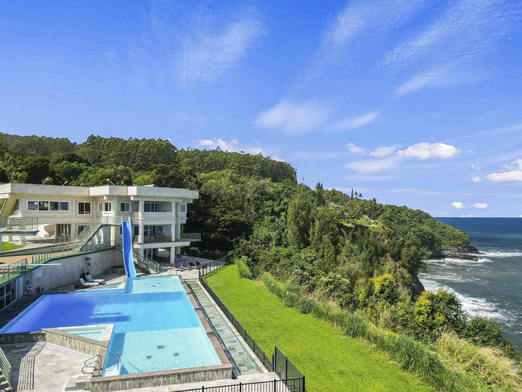 A modern building overlooks a swimming pool, grassy lawn, and ocean.