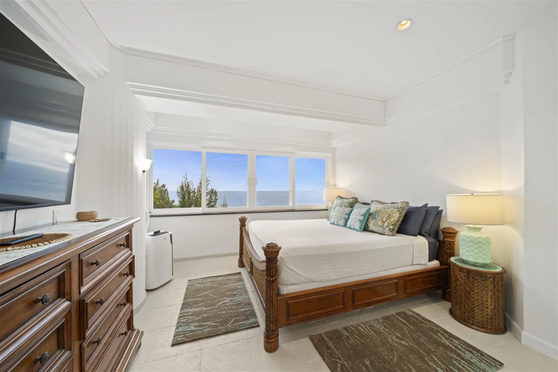 A bright bedroom with a wooden bed, wide windows, and a dresser with a television.