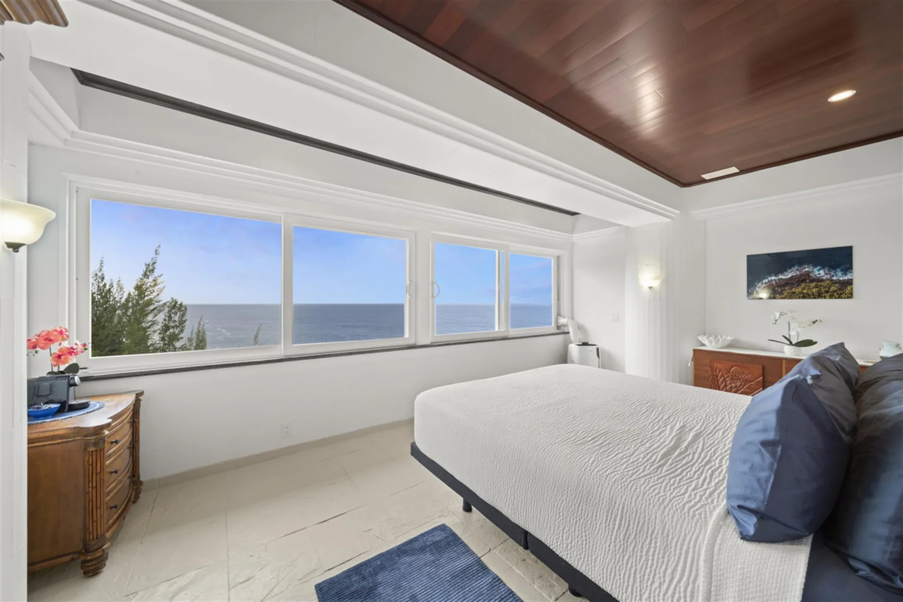 A bright bedroom with a bed beside wide windows overlooking the ocean.