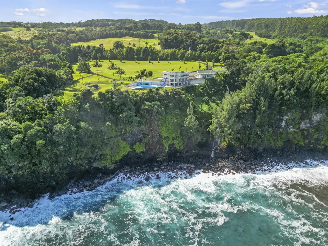 A coastal estate sits above a rocky shoreline, surrounded by dense green hills.