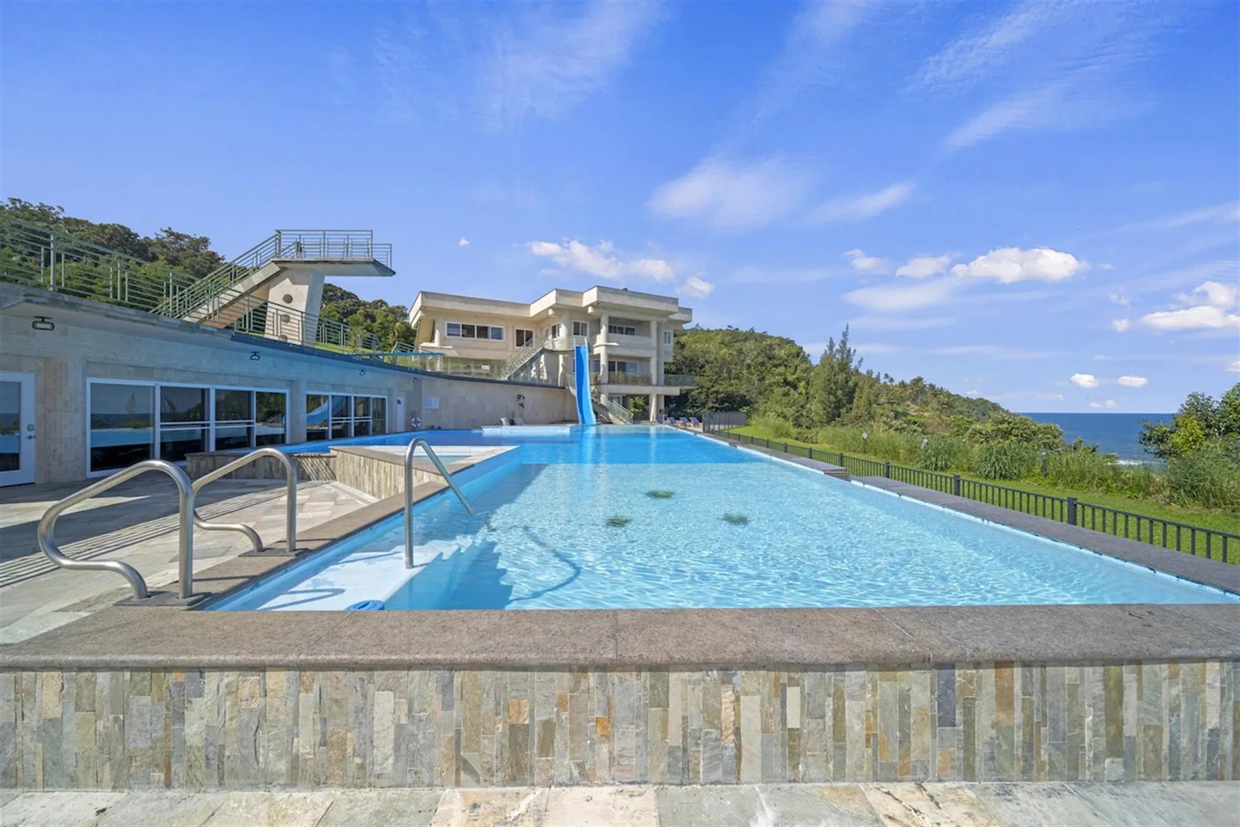 An outdoor swimming pool beside a modern building, with greenery and the sea beyond.