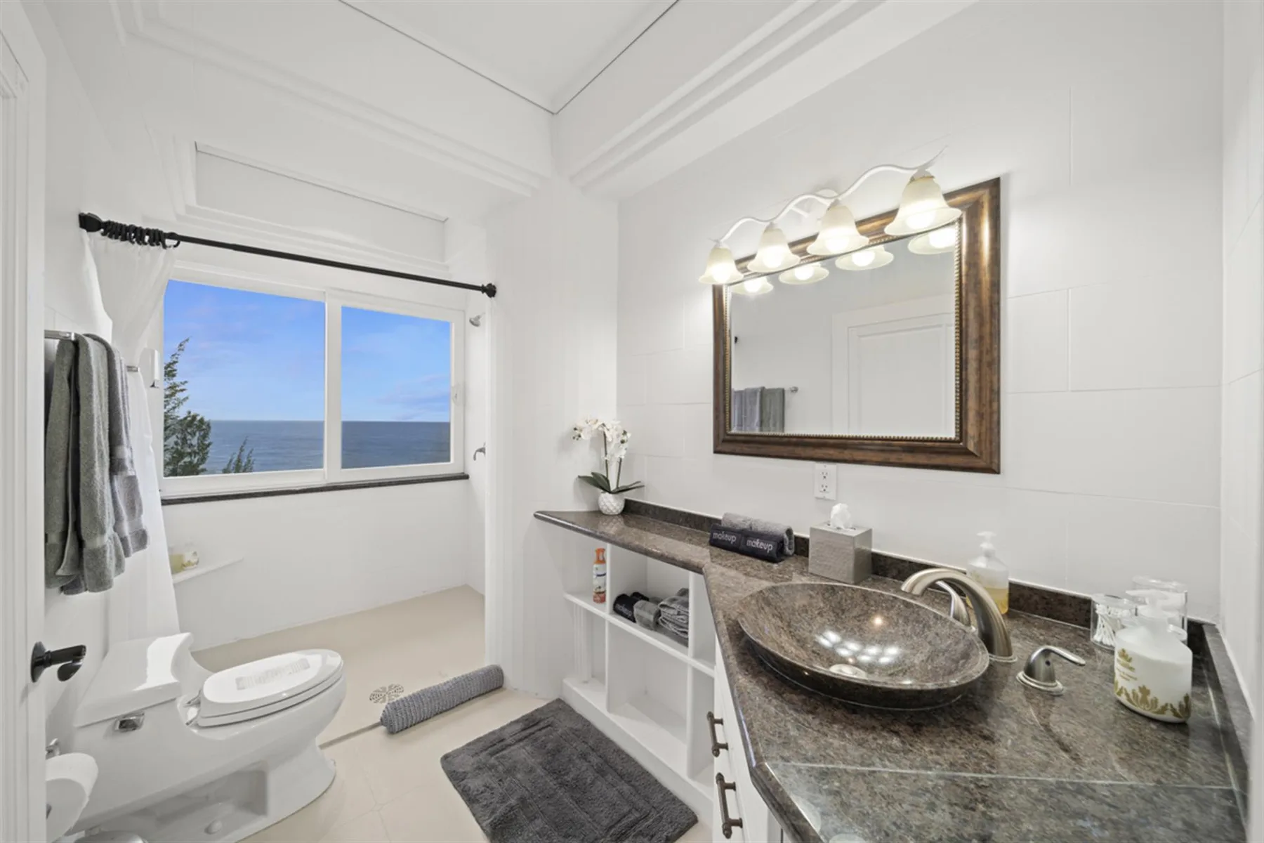 A bright bathroom has a vanity with a mirror beside a window overlooking the ocean.