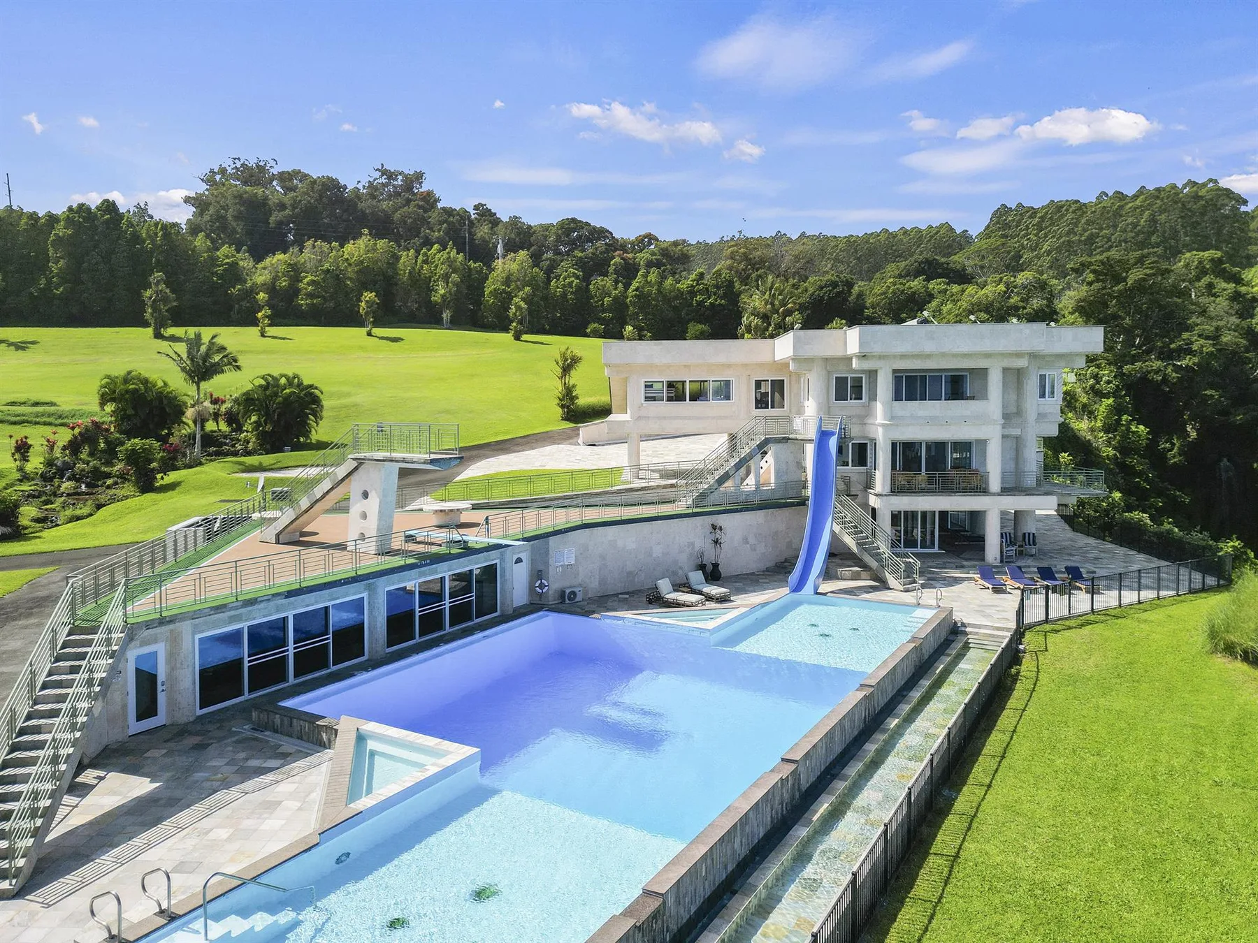 A modern house overlooks a large outdoor pool and surrounding green lawn.
