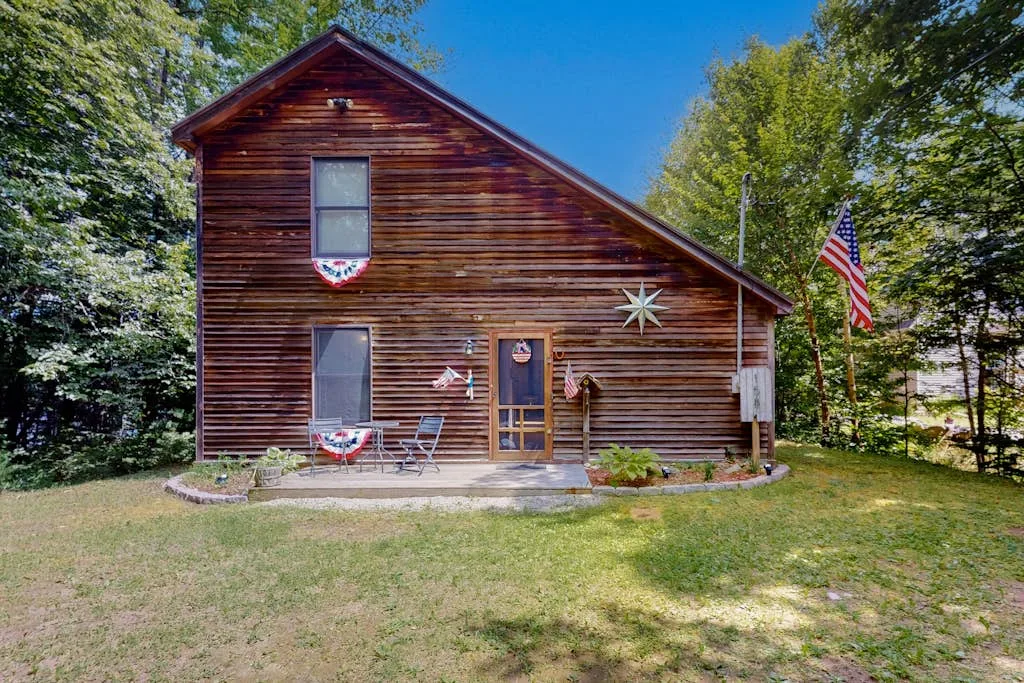 A wooden cabin stands among trees, with a small lawn and an American flag.