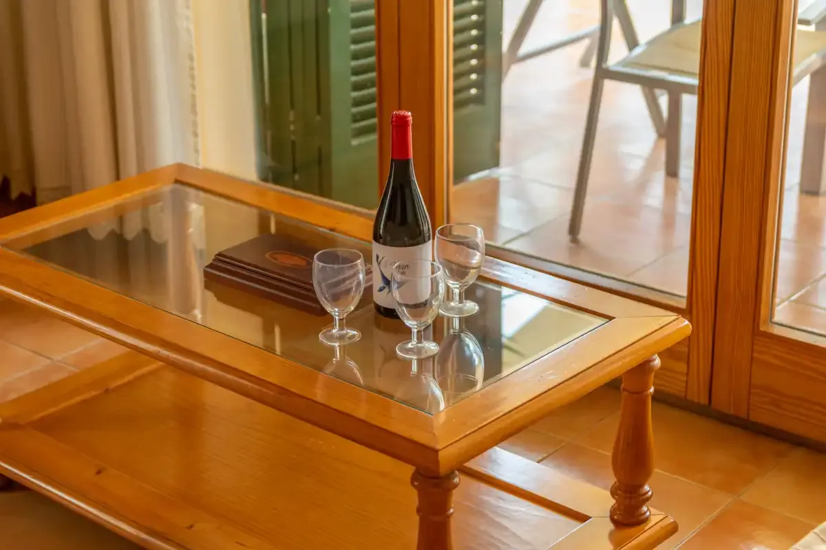 A glass-topped wooden table holds a wine bottle and glasses beside a window.