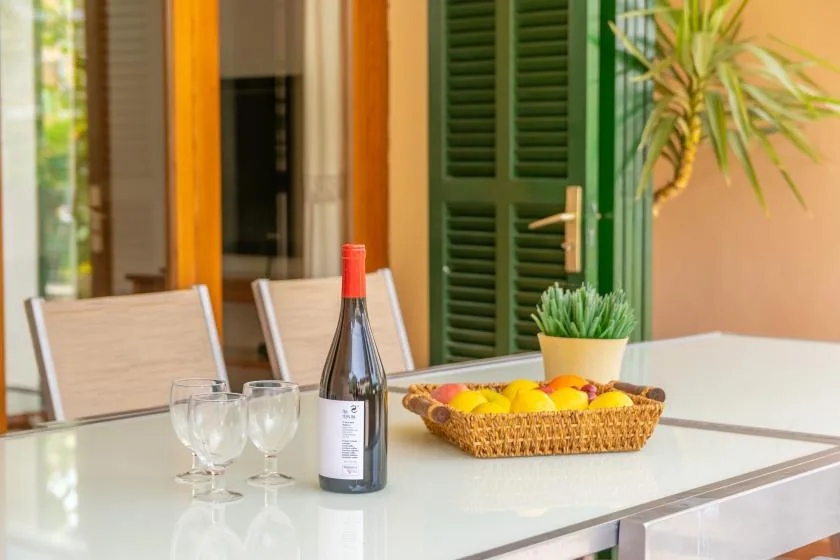 A dining table holds wine glasses and a fruit basket beside green shutters.