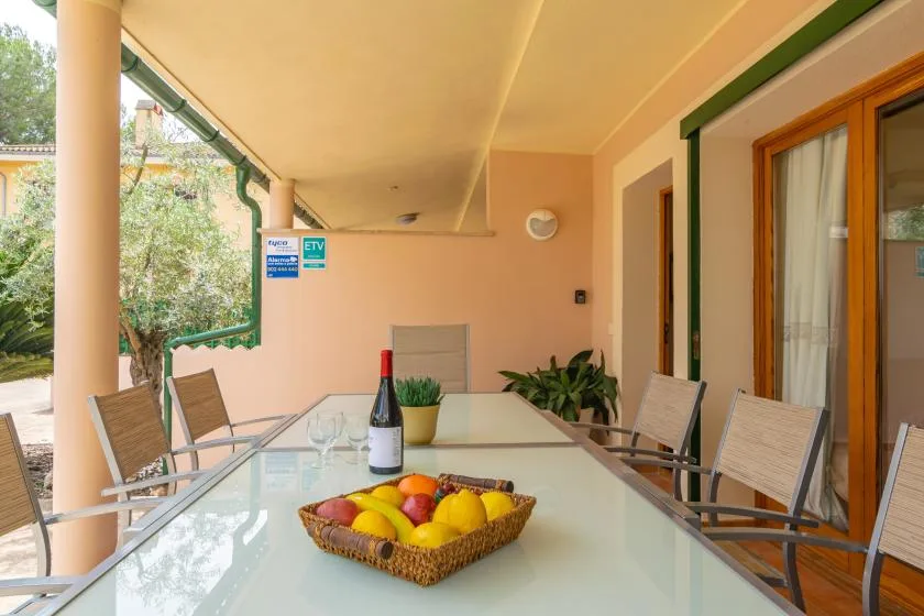 A covered outdoor dining area has a glass table, metal seating, and a fruit basket.