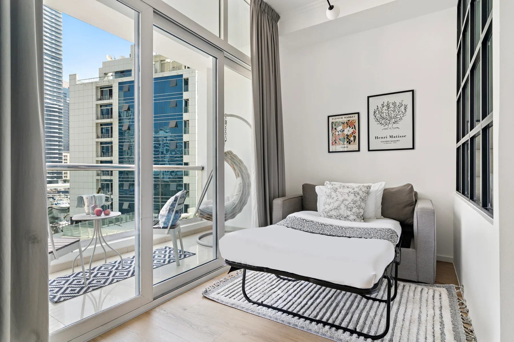 Una habitación luminosa con una cama junto a grandes ventanales que dan a edificios cercanos.