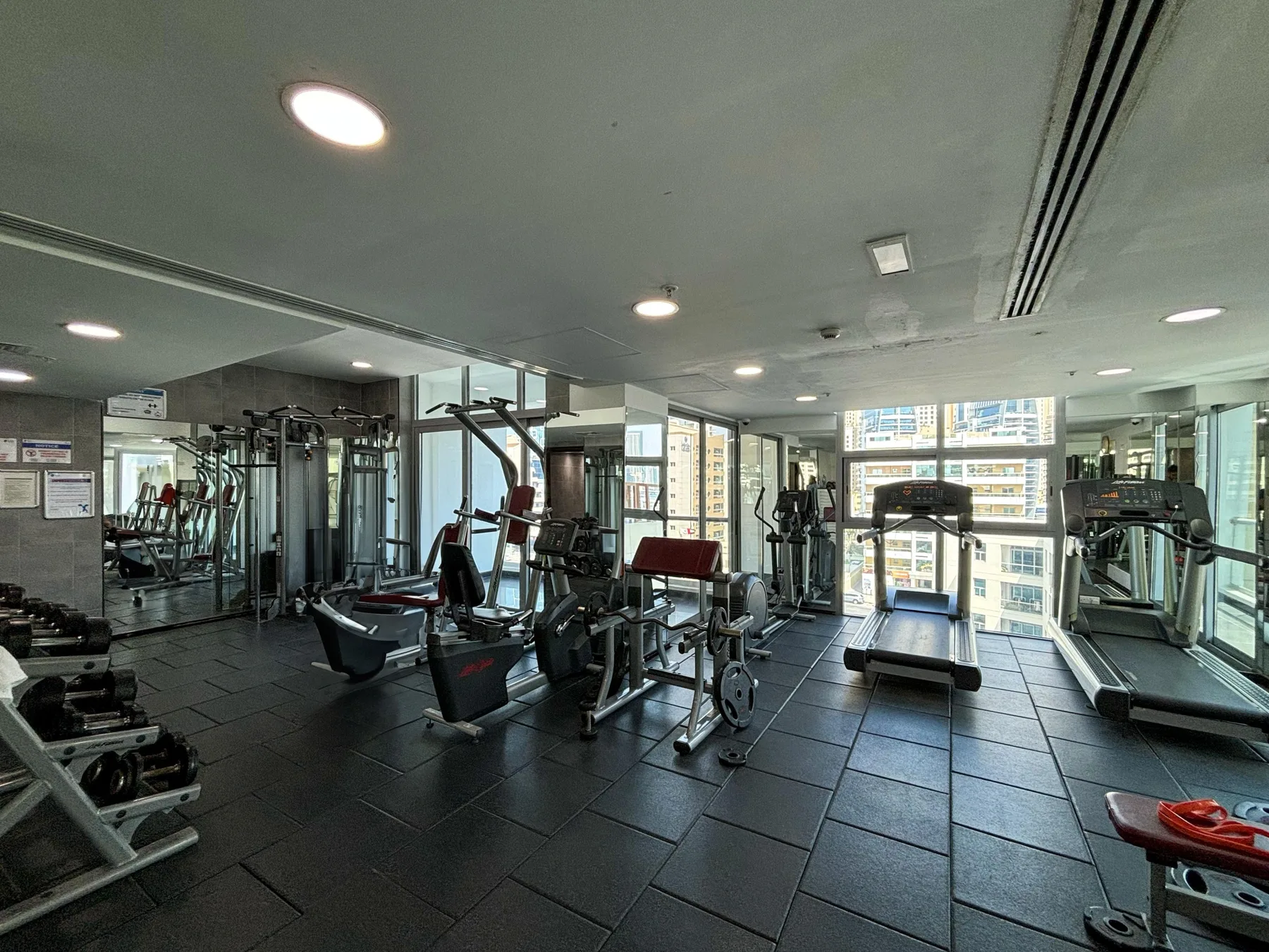 Un gimnasio con máquinas de ejercicio y cintas de correr junto a grandes ventanas.