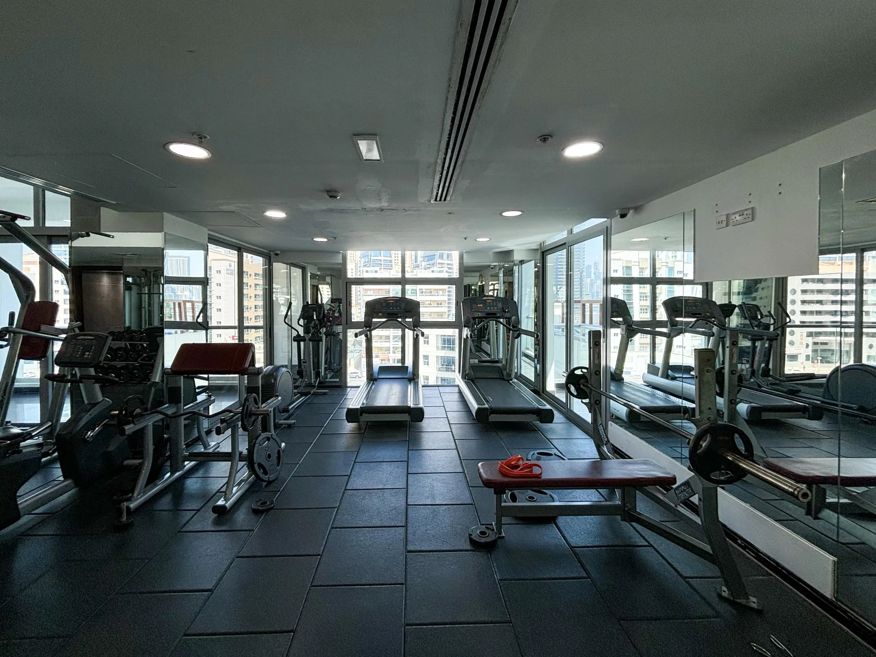 Un gimnasio con cintas de correr y equipo de pesas junto a grandes ventanales.
