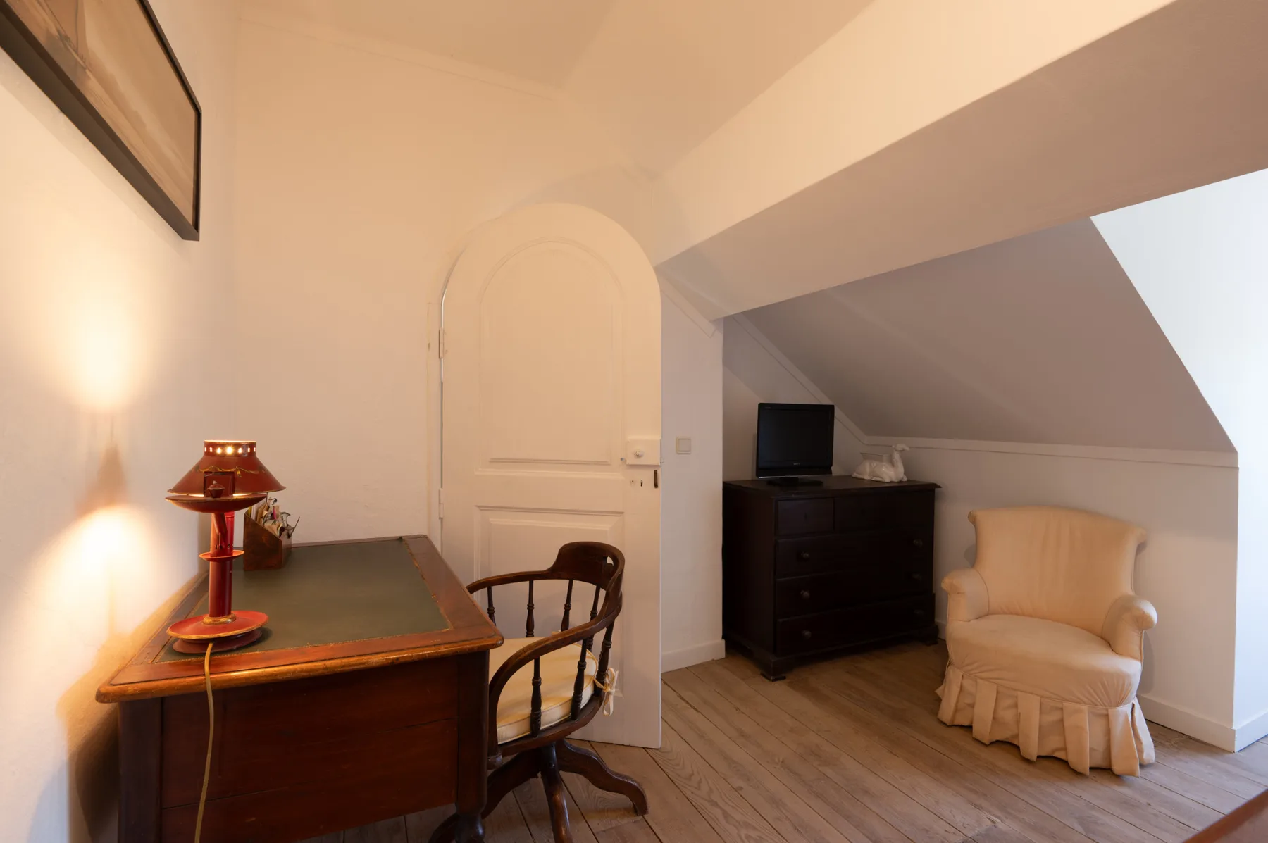 Una habitación abuhardillada amueblada con escritorio, televisor y sillón.