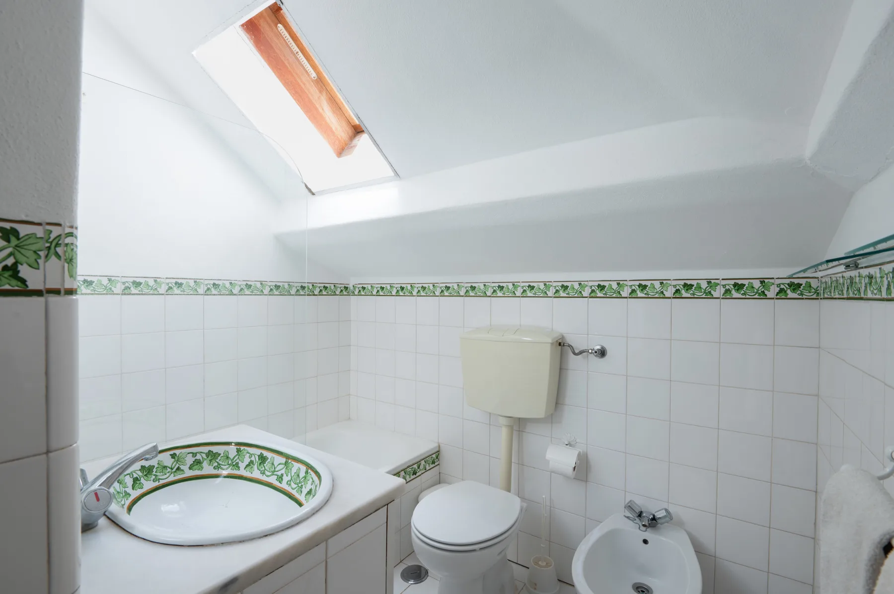 Un baño alicatado con lavabo, inodoro y bidé bajo un techo inclinado.