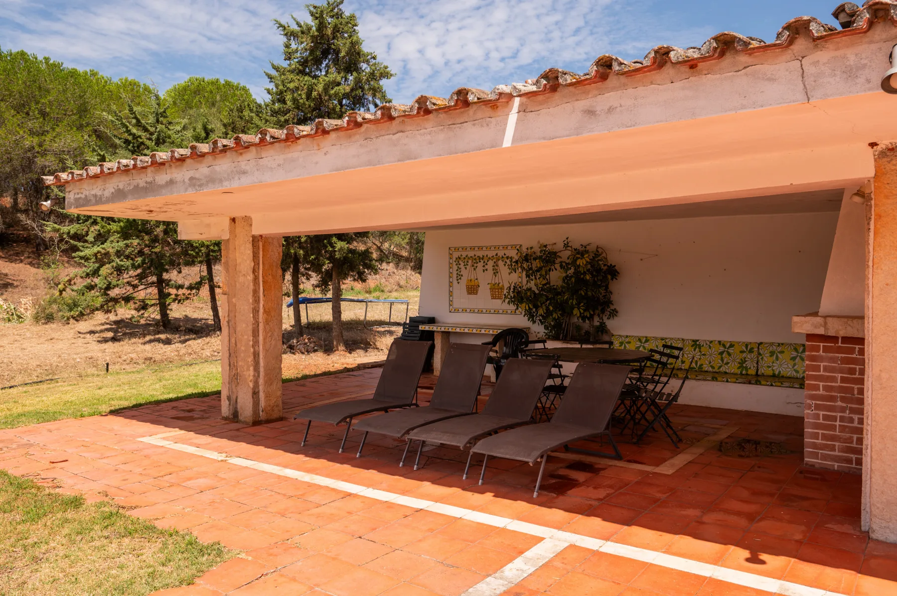 Un patio cubierto con asientos reclinables y suelo de baldosas da a un jardín.
