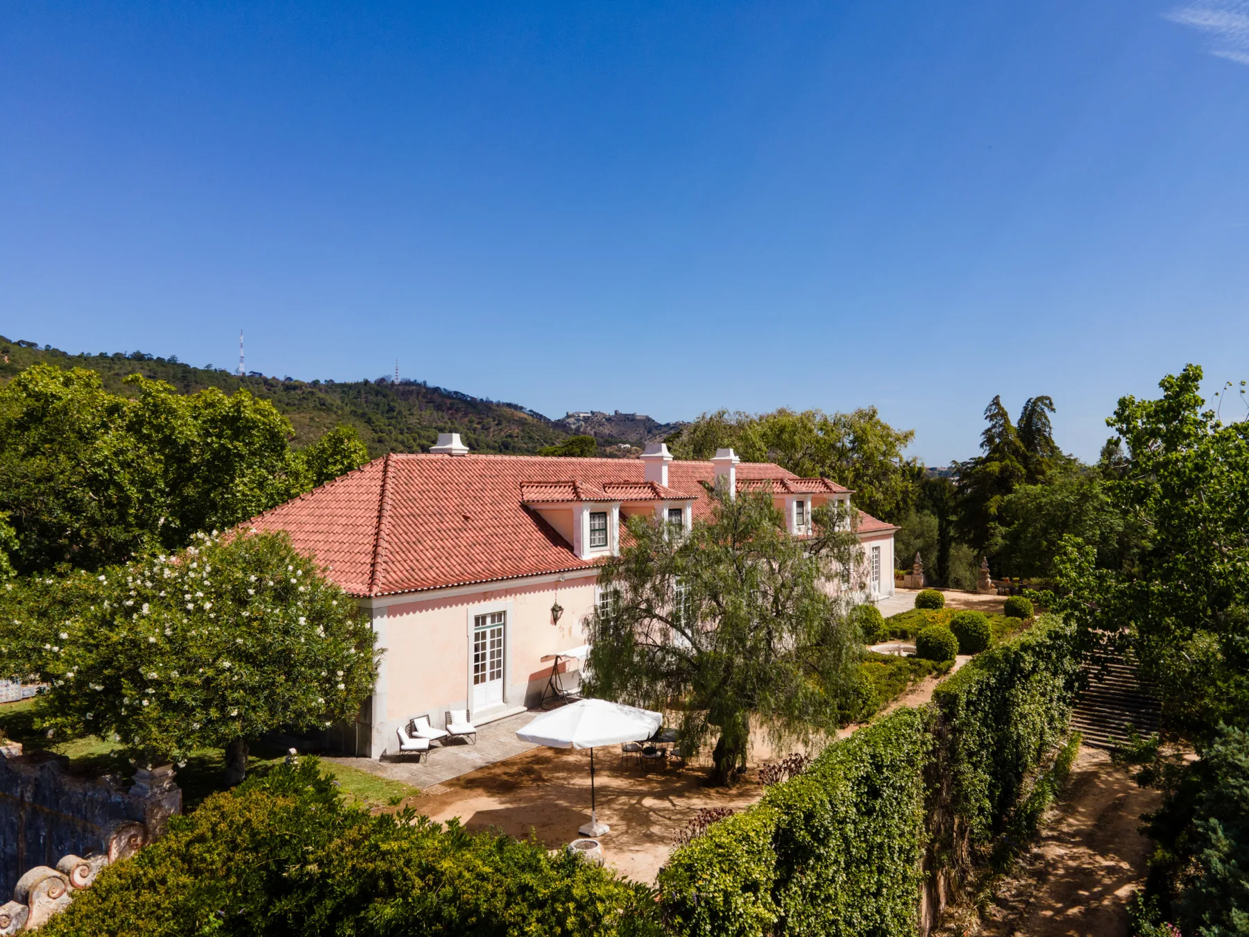 Una casa clara con tejado de tejas rojas está rodeada de jardines y árboles verdes.