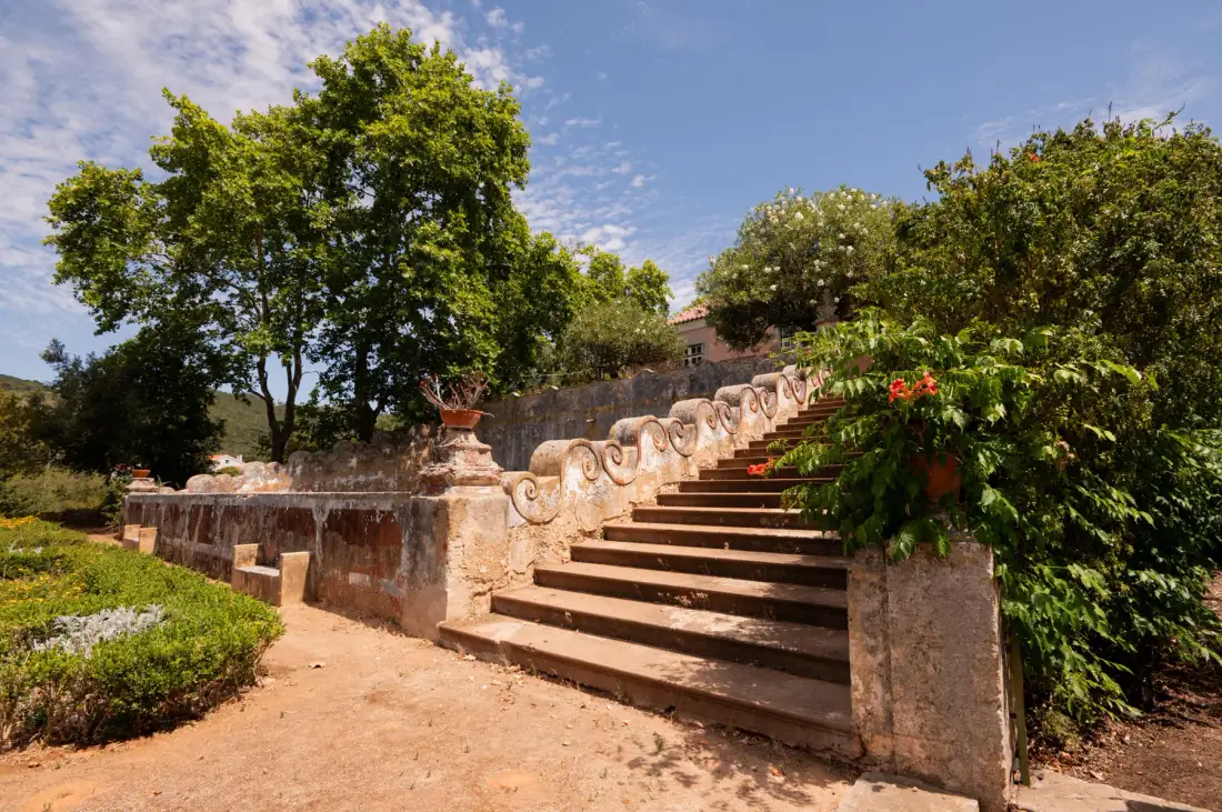 Una escalera de jardín con barandillas de piedra sube entre árboles y plantas con flores.