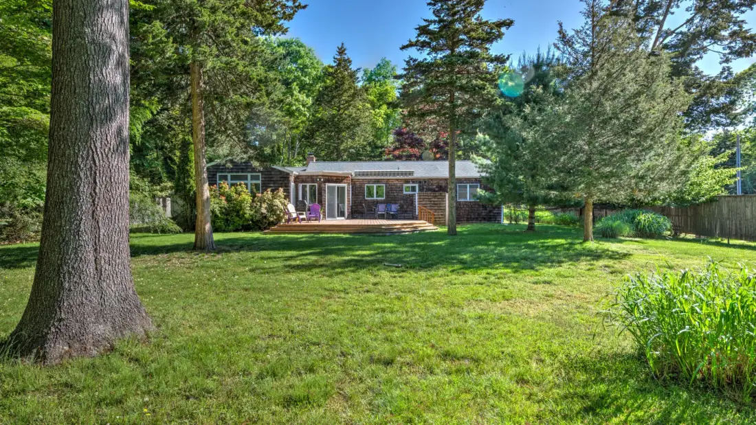 A small house sits beyond a broad lawn, surrounded by mature trees.