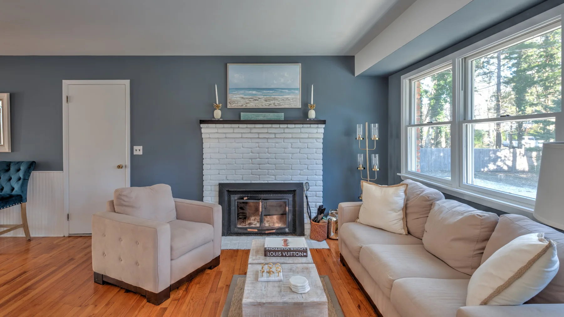 A living room with a fireplace, seating, and large windows overlooking trees.