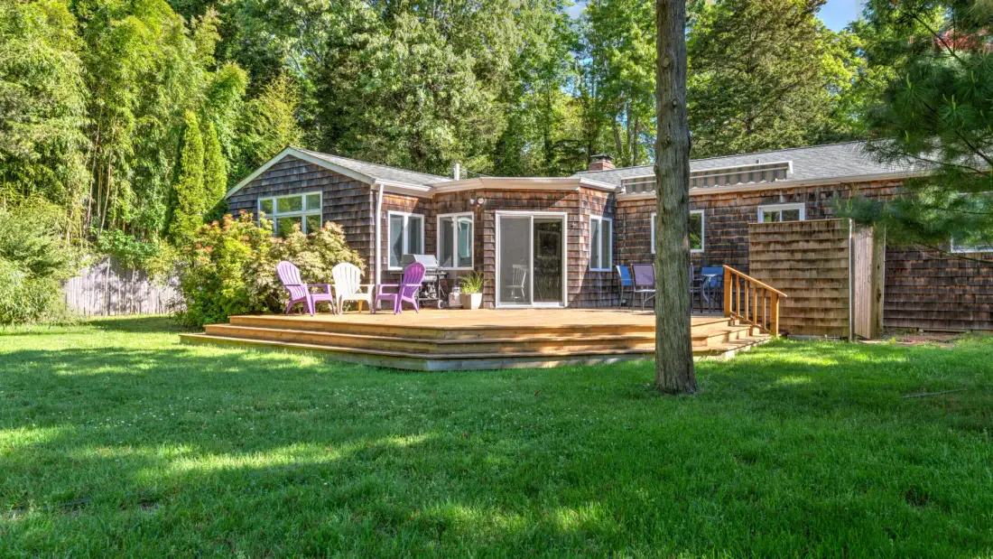 A low, wood-sided house opens onto a raised deck and green lawn.