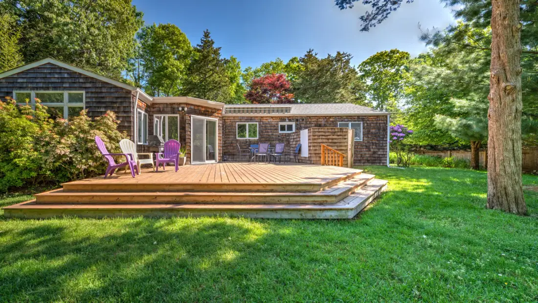 A house with a broad wooden deck opens onto a green lawn.