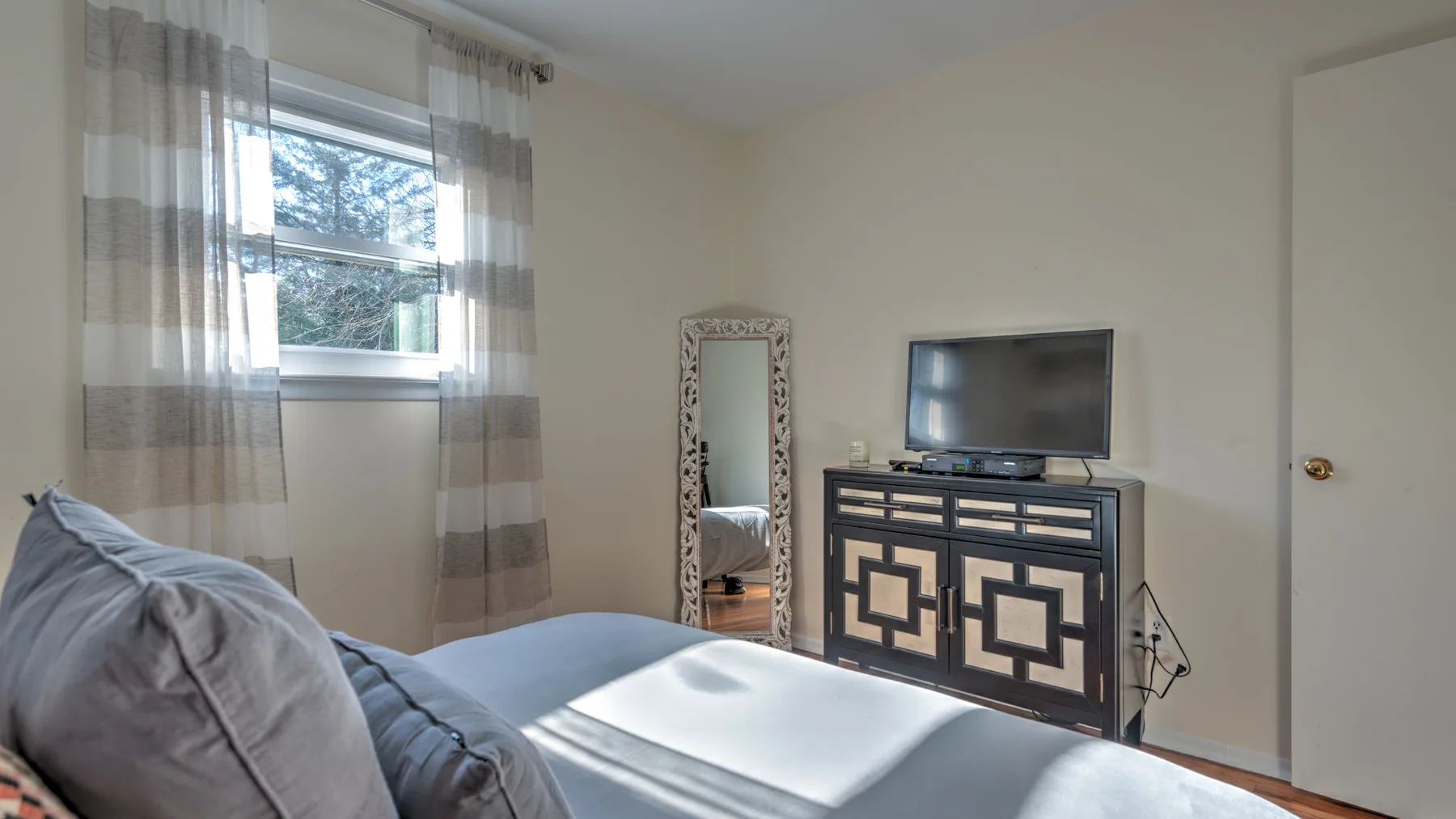 A bedroom with a bed, a television on a cabinet, and a window with sheer curtains.