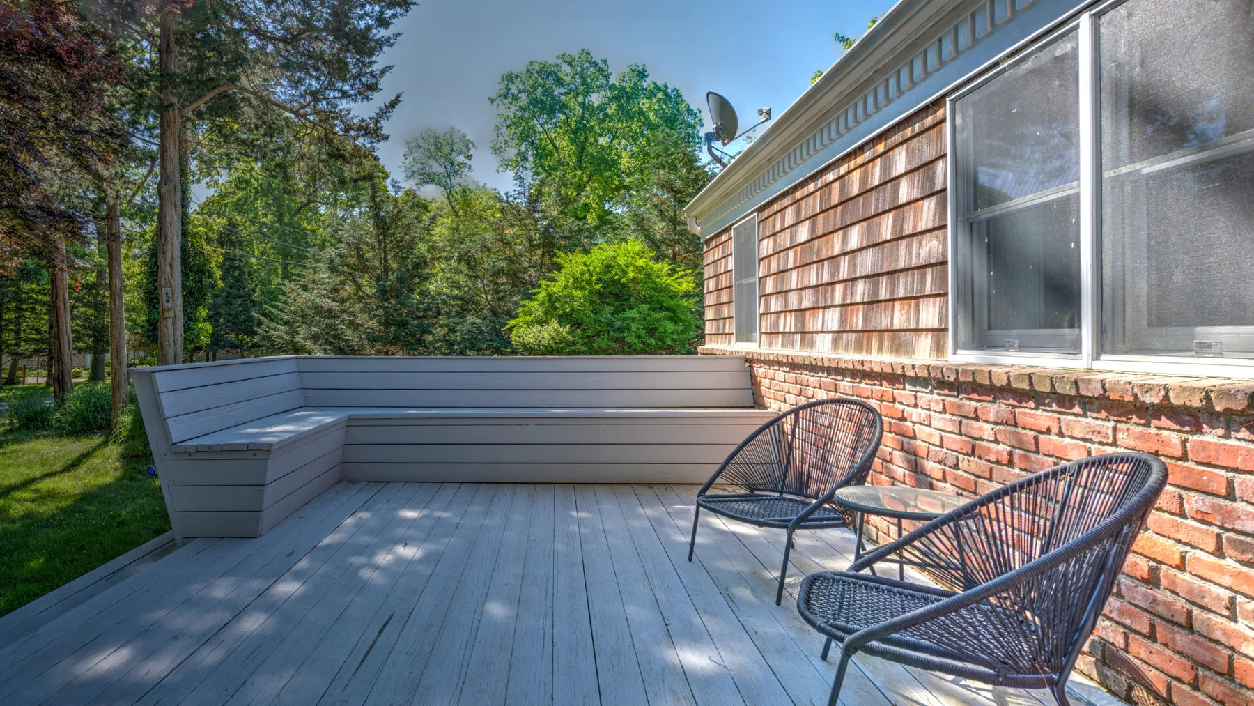 A wooden deck beside a brick-sided house has built-in seating and two outdoor chairs.