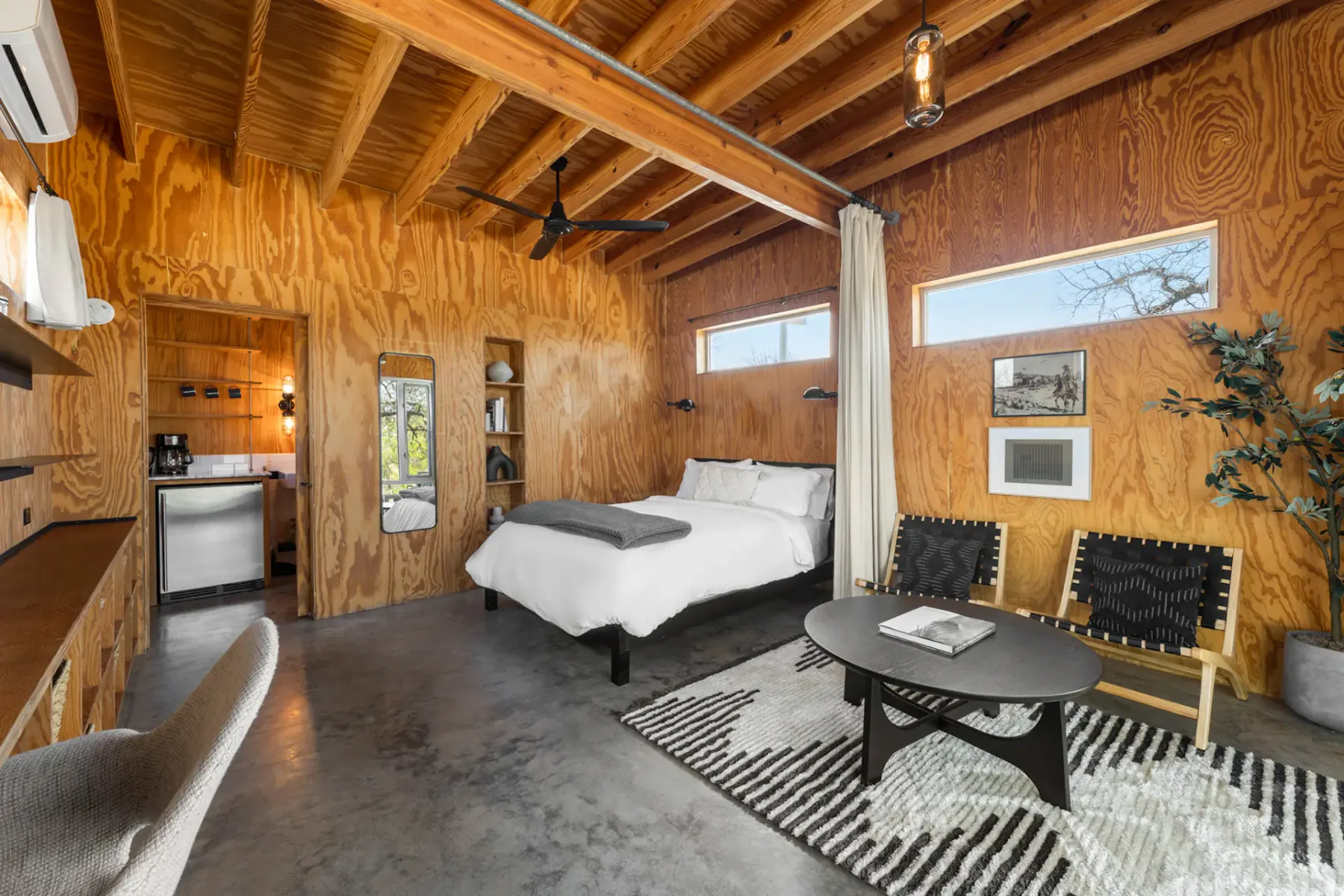 Cozy wooden cabin interior with a bed, seating area, small kitchenette, and modern decor. Natural light streams through narrow windows.