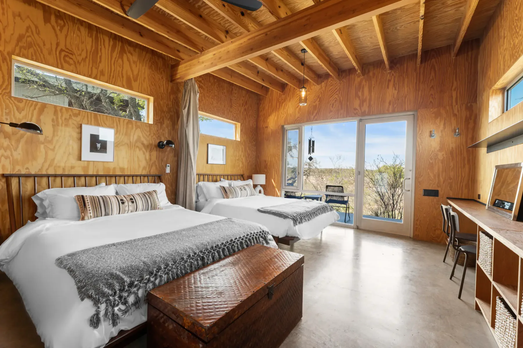Spacious bedroom with two beds, wooden walls and ceiling, large window with scenic view, wicker trunk, and a wooden desk with chair.