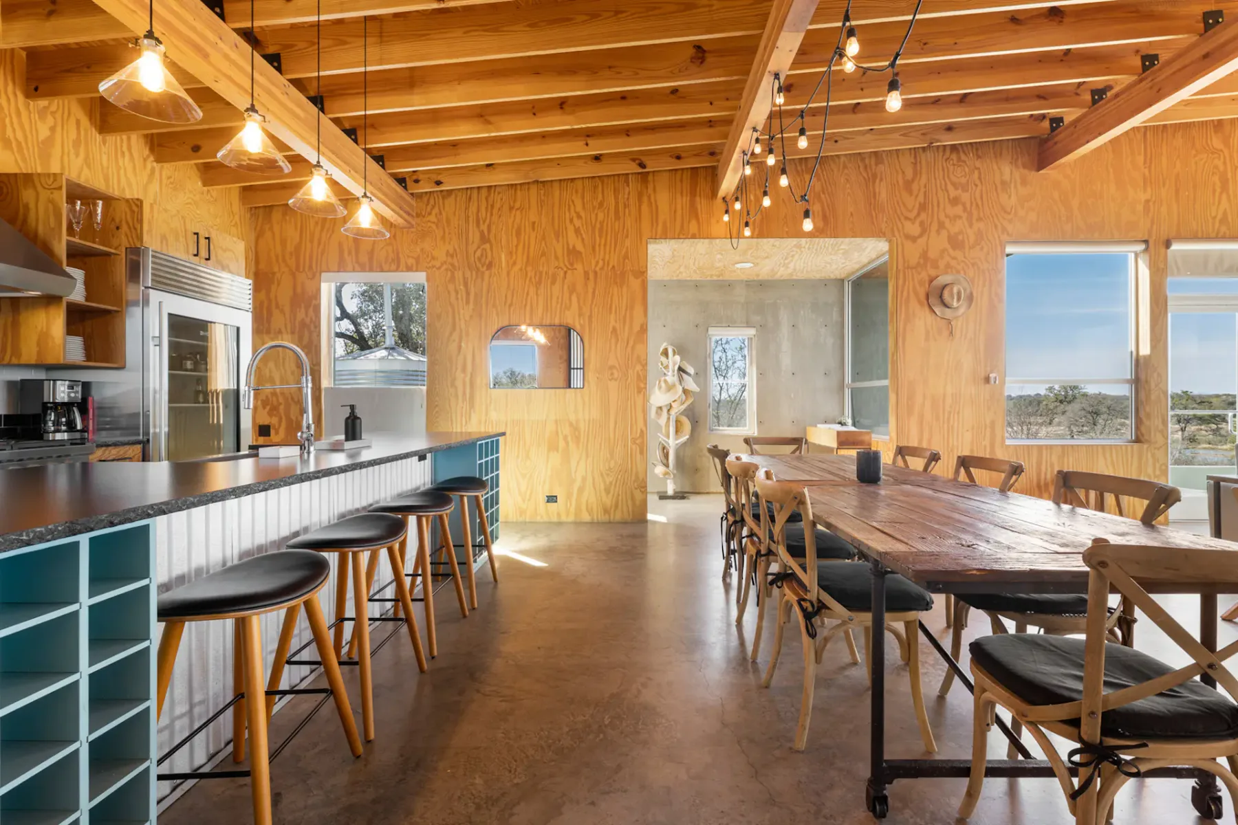 Modern kitchen and dining area with wooden accents, large table, bar stools, hanging lights, and windows offering a scenic view.