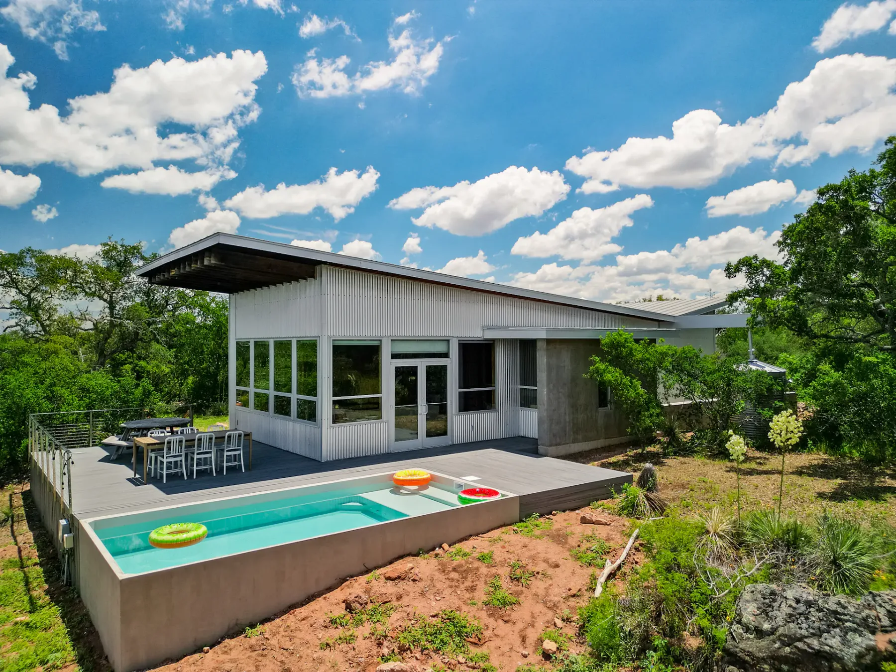 Modern house with large windows and slanted roof, featuring a patio, outdoor dining set, and small pool with inflatables, surrounded by greenery.