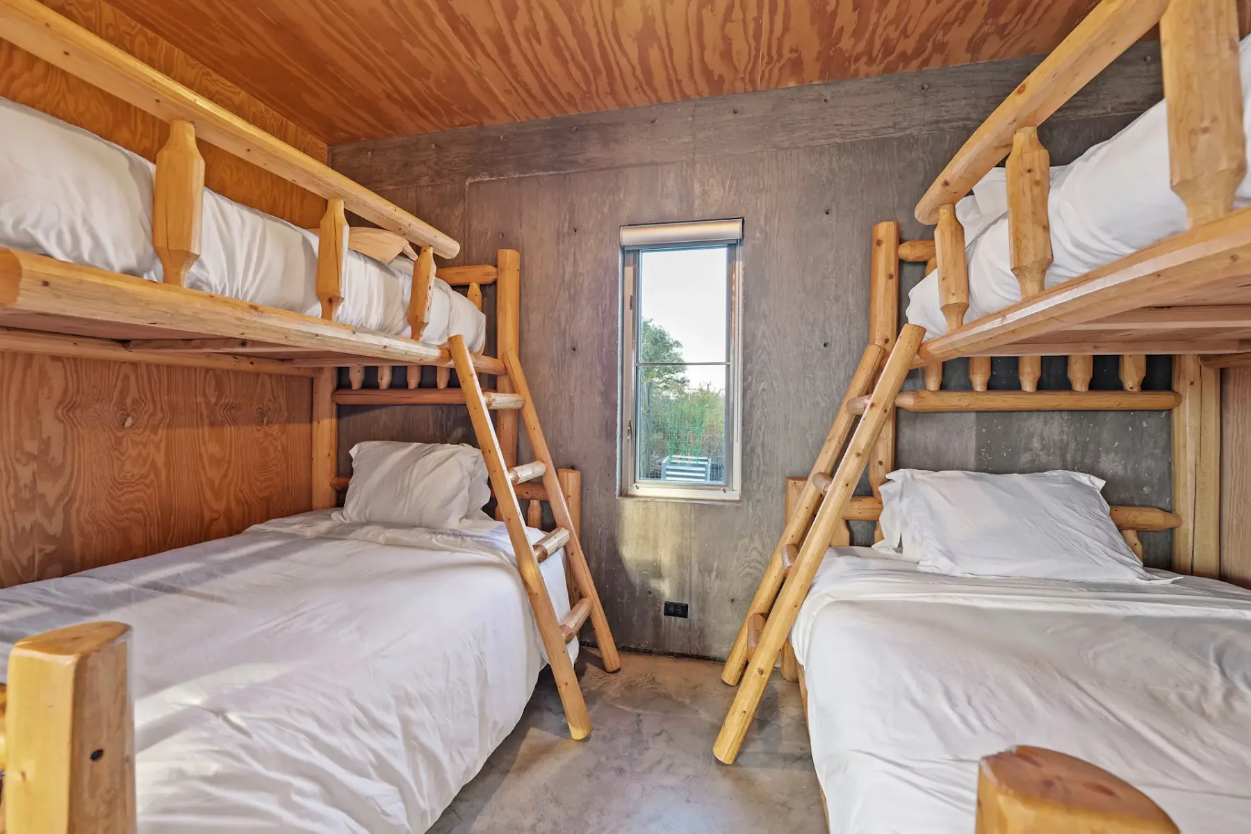 Rustic room with two wooden bunk beds, white bedding, and a central window with a view of trees. Wood-paneled and concrete walls.