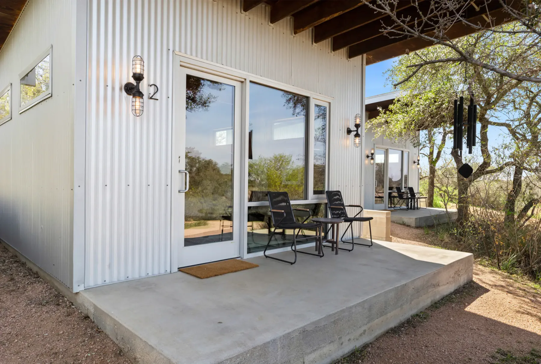Modern metal-clad building with a small patio featuring two chairs and a table. Trees and outdoor lighting enhance the serene setting.