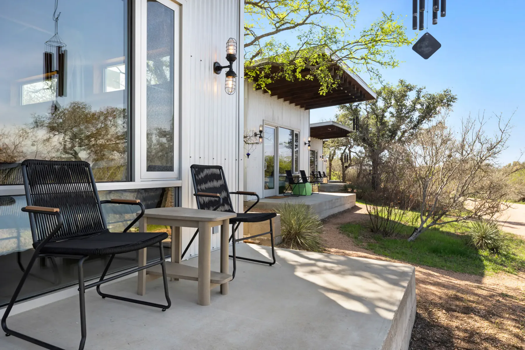 Patio area with two black chairs and a wooden table, adjacent to a modern white building and overlooking a natural landscape with trees.