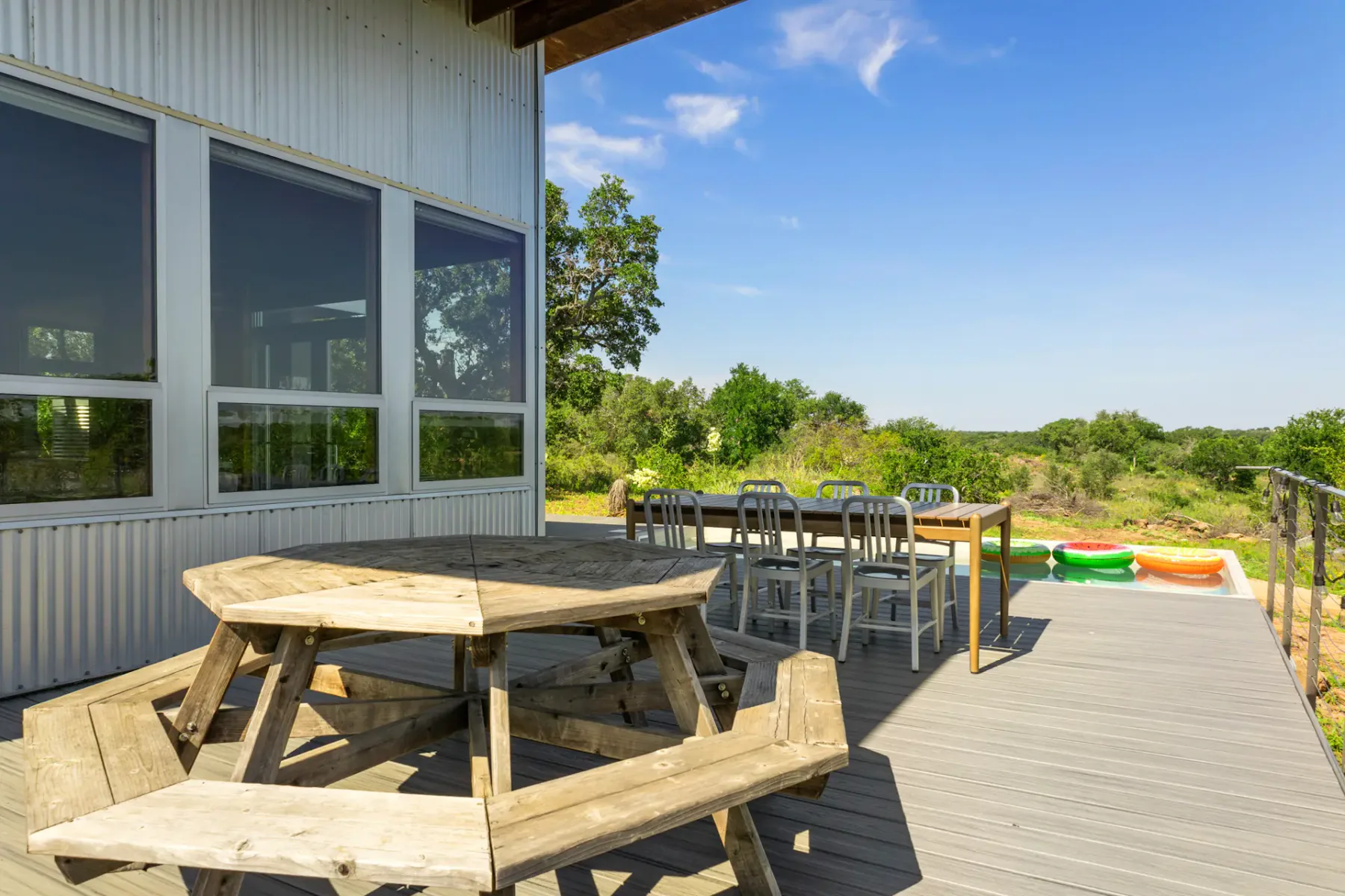 Wooden picnic table on a deck near a modern building, with additional seating and a view of greenery under a clear blue sky.