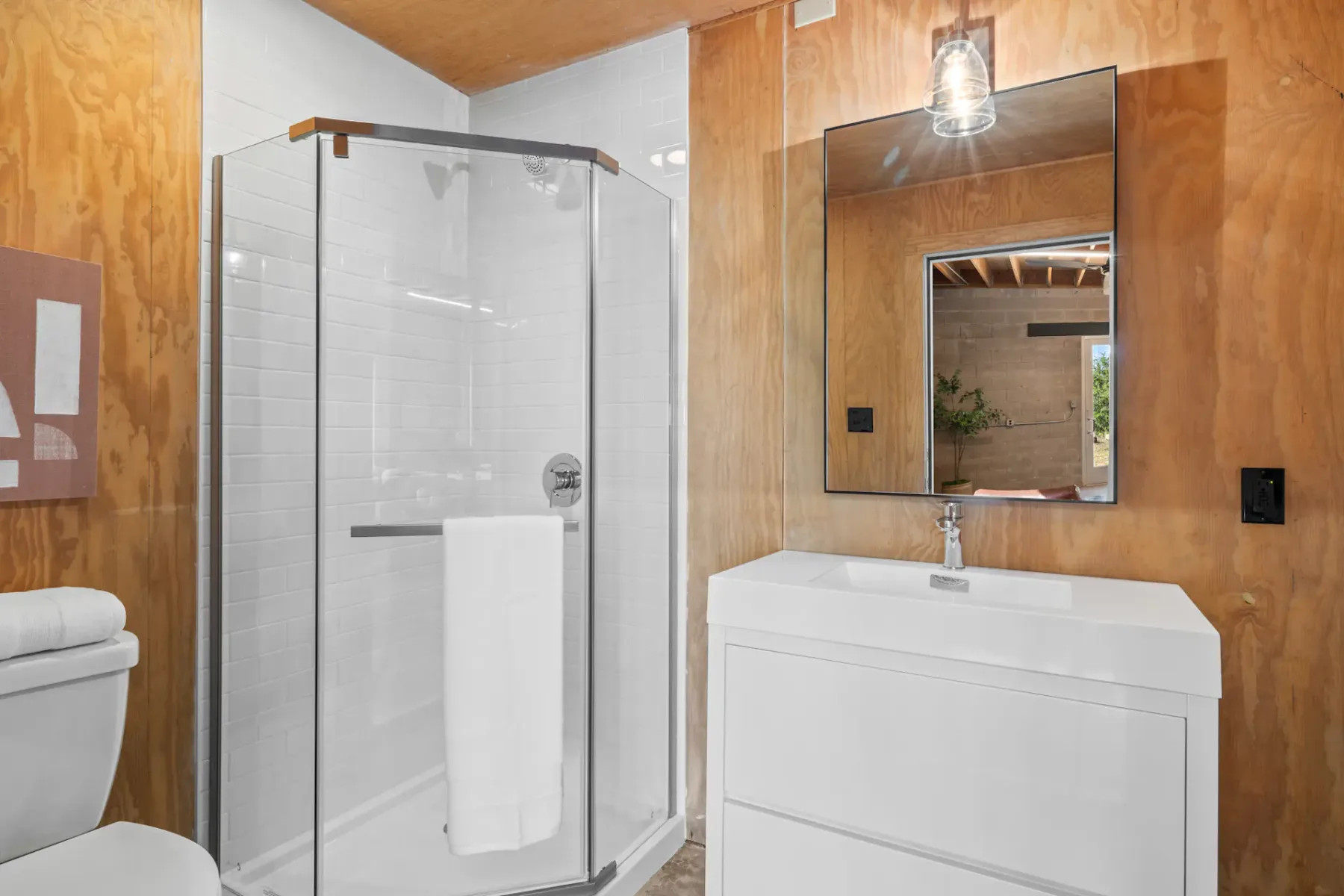 Modern bathroom with a glass shower, white vanity, square mirror, and wood-paneled walls. A towel is hanging inside the shower.