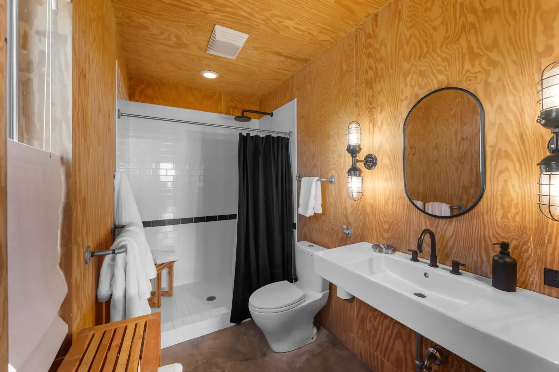 A modern bathroom with wood paneling, a shower with a black curtain, a round mirror above a white sink, and industrial wall lights.