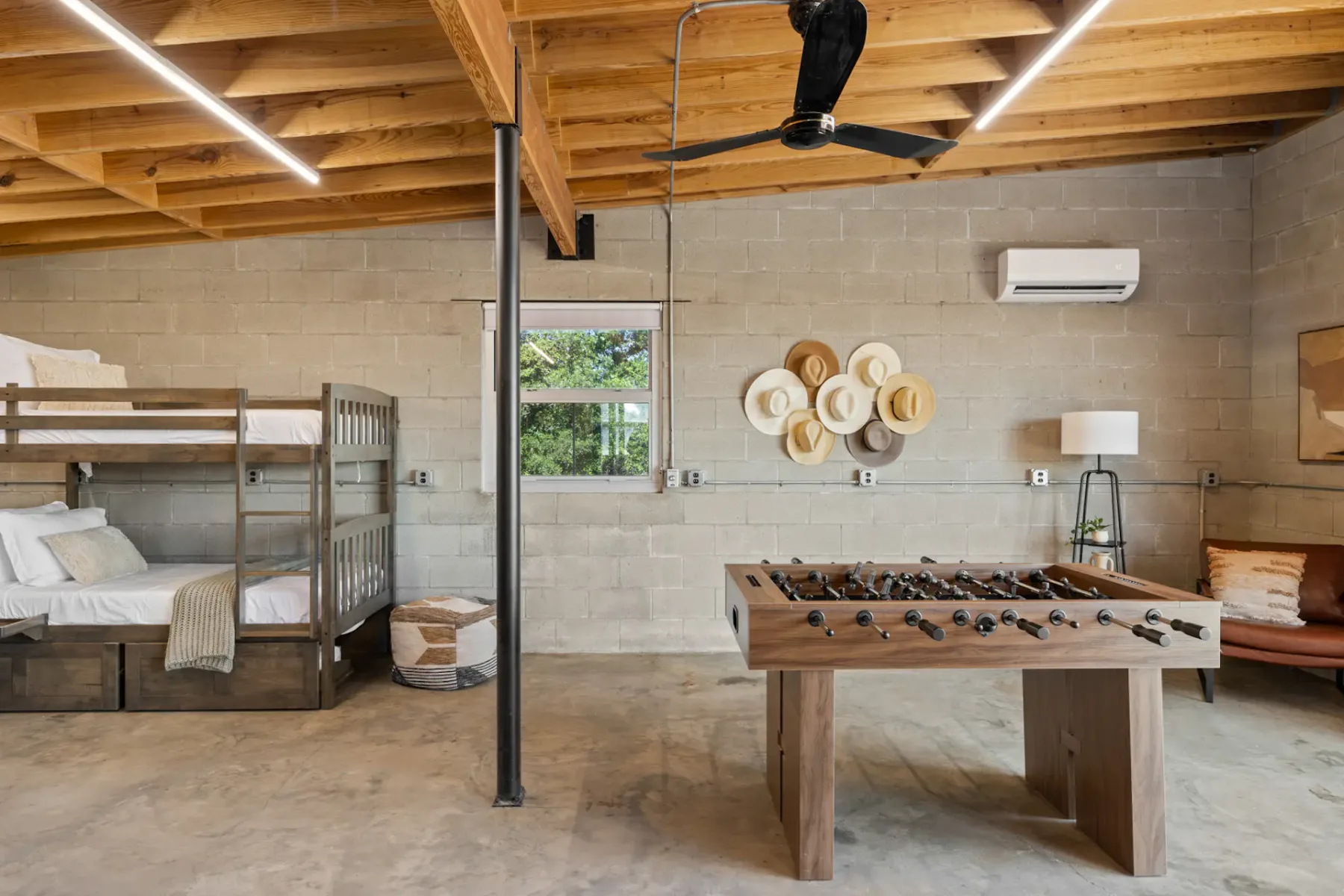 Spacious room with a foosball table, bunk beds, hats on the wall, ceiling fan, and a large window showing greenery outside.