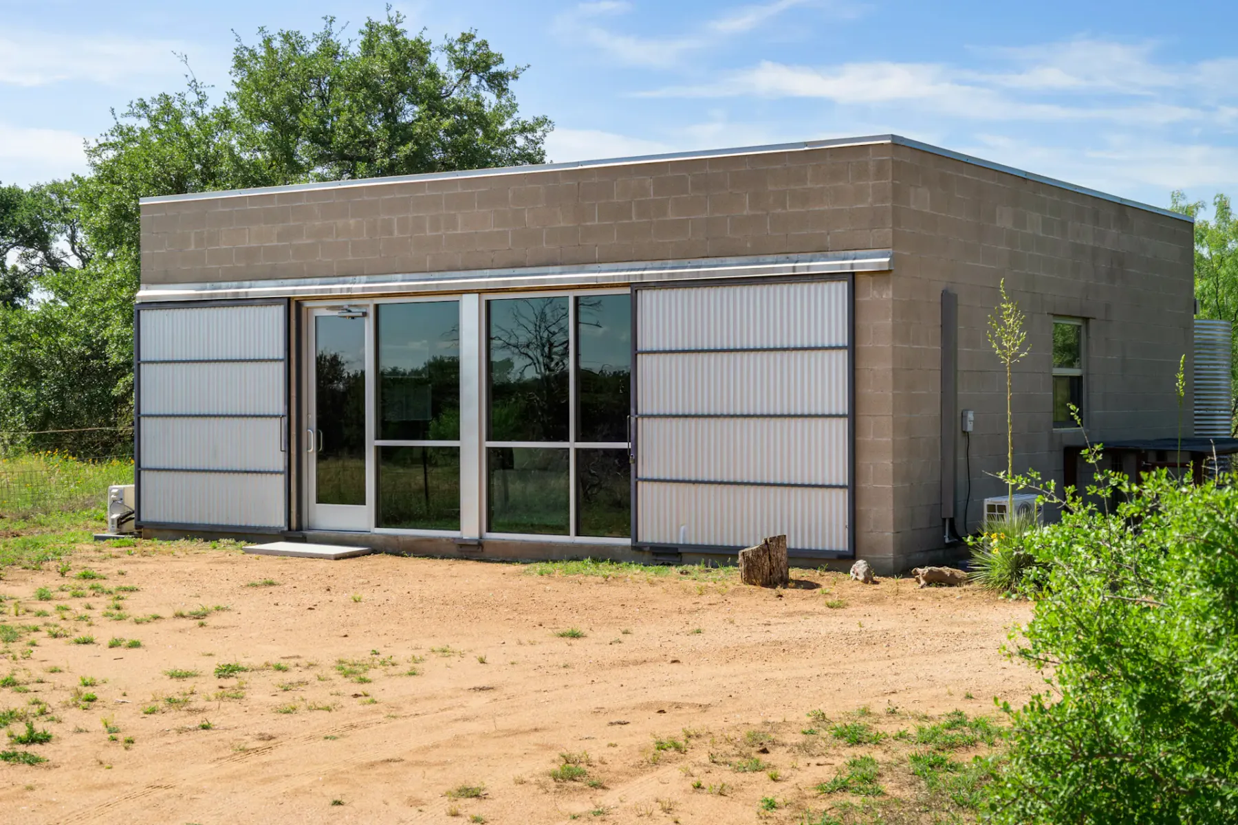 A modern, cube-shaped concrete building with large glass doors and windows surrounded by a dirt yard and sparse greenery.