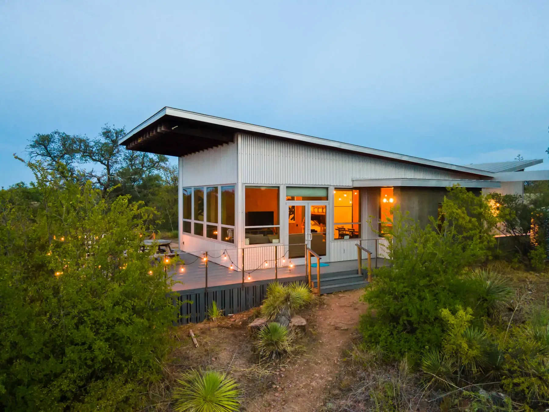 Modern house with a slanted roof, large windows, and a wooden deck, set among greenery at dusk with warm interior lights visible.