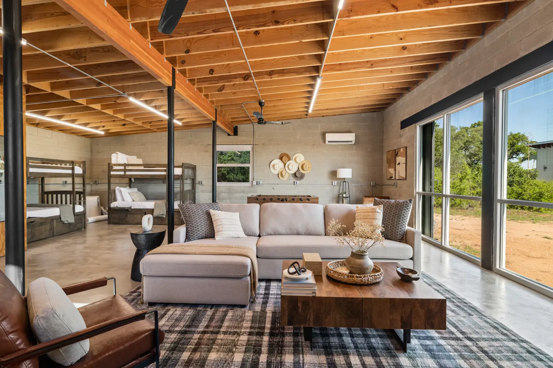 Spacious living area with modern decor, exposed wooden beams, large windows, sectional sofa, and a view of the bunk beds in the background.