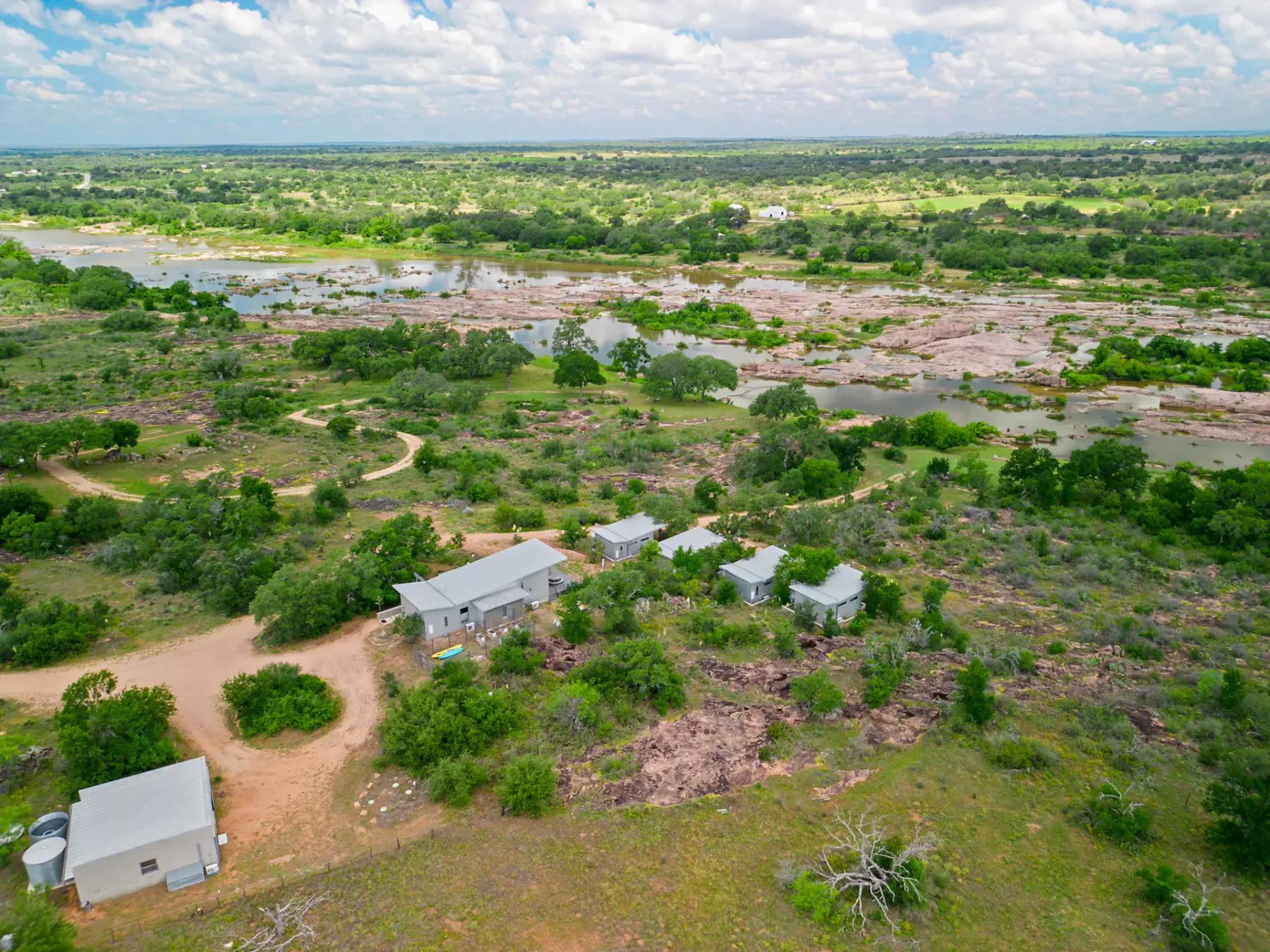 Aerial view of a rural landscape with scattered buildings, green vegetation, dirt paths, and a cloudy sky.